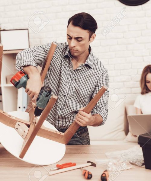 Man with Drill in Hands Repairing Chair in Room.